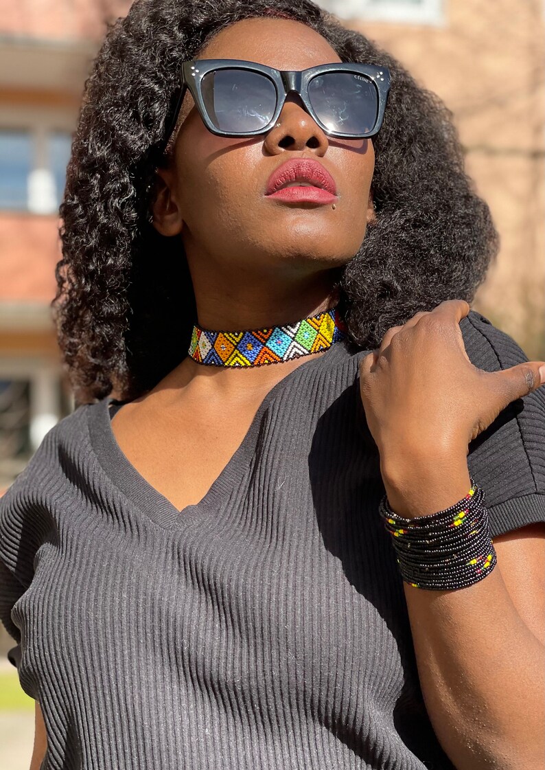 May include: A beaded choker necklace and bracelet set. The choker features a geometric pattern in blue, yellow, white, and orange beads. The bracelet is black with yellow and red accents. The person is wearing a black V-neck top.