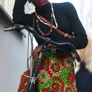 May include: A person wearing a black turtleneck and a red hat, leaning on a bicycle. The skirt features a vibrant floral pattern in red, green, and white. Accessories include layered necklaces and large square earrings.