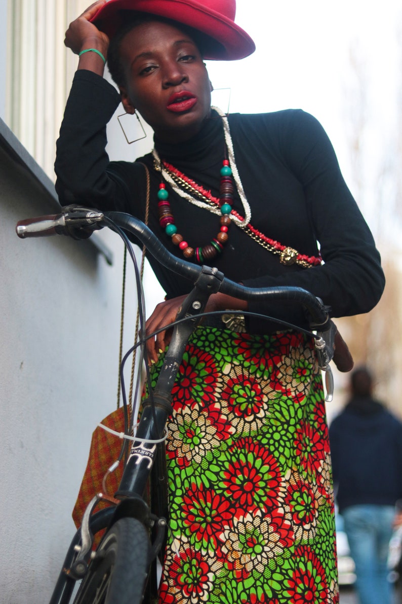 May include: A person wearing a black turtleneck and a red hat, leaning on a bicycle. The skirt features a vibrant floral pattern in red, green, and white. Accessories include layered necklaces and large square earrings.