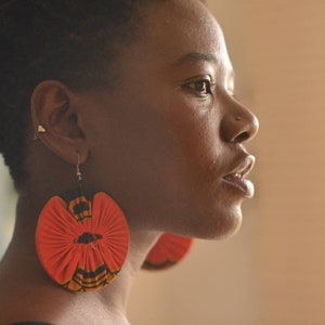 May include: A woman with short black hair wearing large, round, red and black fabric earrings.
