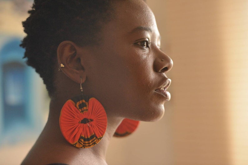 May include: A woman with short black hair wearing large, round, red and black fabric earrings.