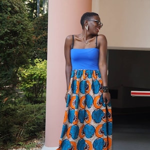 K&ouml;nnte beinhalten: Eine Frau tr&auml;gt ein blaues Oberteil mit schwarzen Tr&auml;gern und einen langen, gemusterten Rock. Der Rock hat ein lebendiges Design mit orangefarbenen, blauen und wei&szlig;en kreisf&ouml;rmigen Mustern. Sie tr&auml;gt eine Brille und Schmuck.
