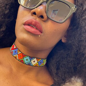 May include: A vibrant beaded choker necklace with a geometric design. The necklace incorporates red, orange, blue, green, and white beads. The person in the image is wearing black sunglasses.