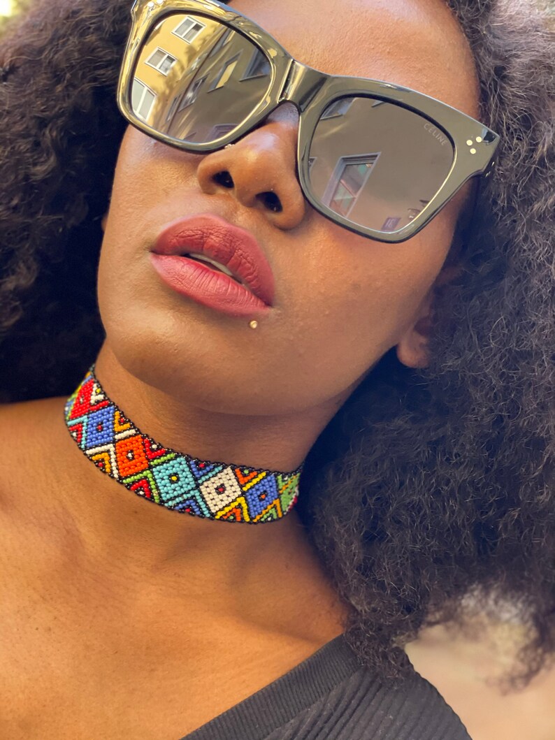May include: A vibrant beaded choker necklace with a geometric design. The necklace incorporates red, orange, blue, green, and white beads. The person in the image is wearing black sunglasses.