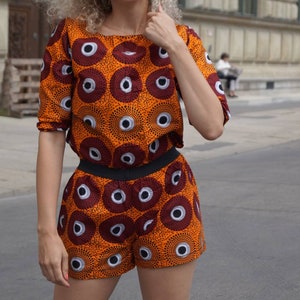 May include: An orange two-piece outfit with a repeating pattern of dark red and white circles. The top is a short-sleeved crop top, and the shorts have a black waistband. The outfit is made of a patterned fabric.