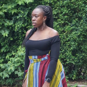 May include: A woman in a black off-the-shoulder top and a wrap skirt with a colourful striped pattern in red, yellow, and blue. She has long black braids and is wearing a watch. The skirt is wrapped around the waist.