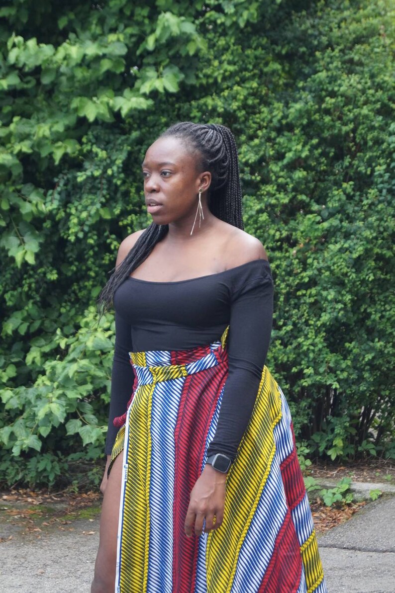May include: A woman in a black off-the-shoulder top and a wrap skirt with a colourful striped pattern in red, yellow, and blue. She has long black braids and is wearing a watch. The skirt is wrapped around the waist.