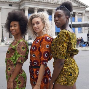 May include: Three people wearing matching short-sleeved romper outfits with vibrant, circular patterns in shades of green, orange, and yellow. The outfits have a high neckline and are paired with shorts. The background features a large building with columns.