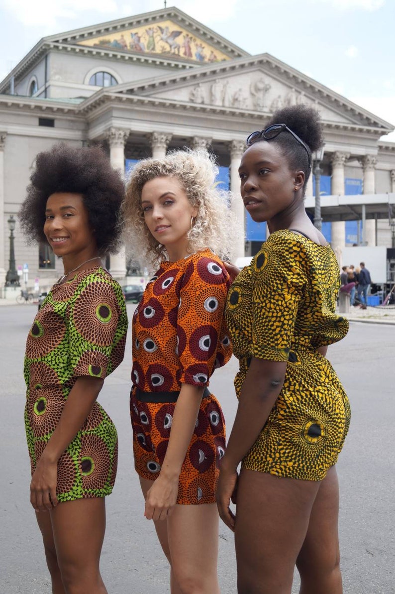 May include: Three people wearing matching short-sleeved romper outfits with vibrant, circular patterns in shades of green, orange, and yellow. The outfits have a high neckline and are paired with shorts. The background features a large building with columns.