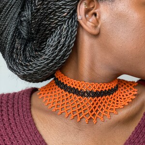 Może przedstawiać: Pomarańczowy i czarny naszyjnik typu choker z koralik&oacute;w. Naszyjnik ma geometryczny wz&oacute;r z pomarańczowymi koralikami tworzącymi wz&oacute;r w kształcie rombu i czarną opaską z koralik&oacute;w na g&oacute;rze. Naszyjnik jest noszony przez osobę o ciemnej sk&oacute;rze i czarnych, zaplecionych włosach.