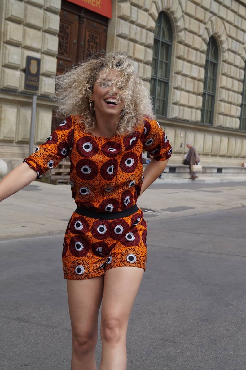 May include: An orange and brown two-piece outfit with a repeating circular pattern. The top has short sleeves and a crew neck, and the shorts have a black waistband. The outfit is worn outdoors.