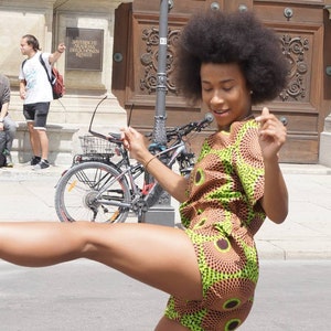 May include: A woman wearing a short-sleeved romper with a brown and green circular pattern. The woman is outside on a sunny day, with a bicycle and building in the background. She is smiling and appears to be dancing.