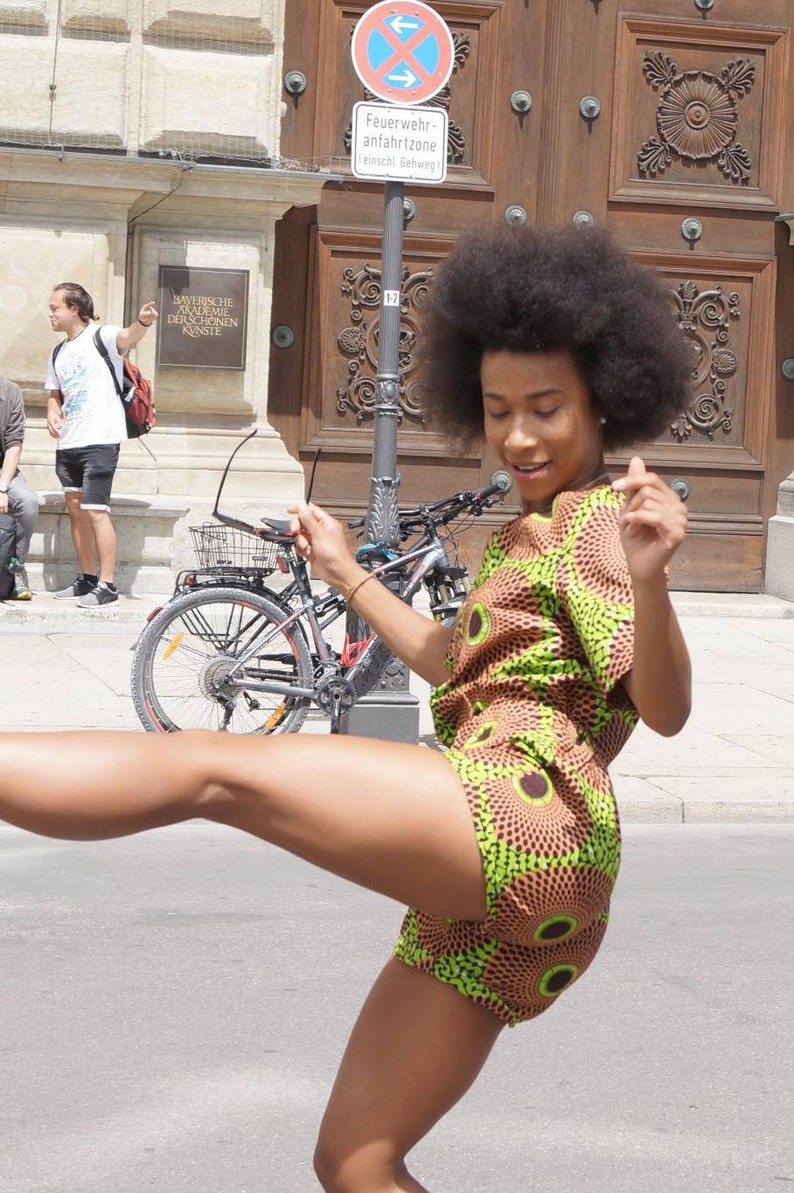 May include: A woman wearing a short-sleeved romper with a brown and green circular pattern. The woman is outside on a sunny day, with a bicycle and building in the background. She is smiling and appears to be dancing.
