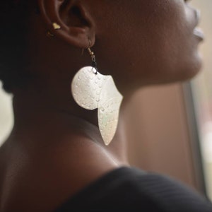 May include: A silver leaf-shaped earring with a textured surface. The earring is hanging from a person's ear.