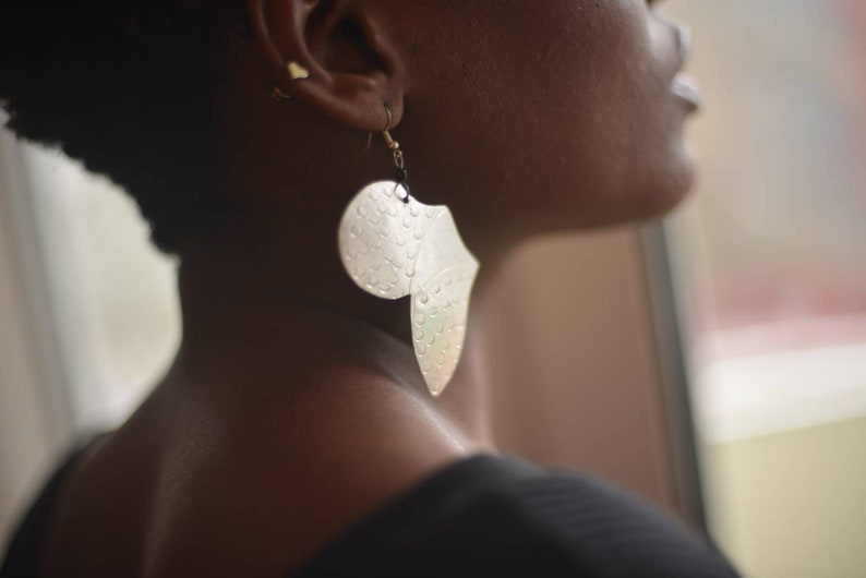 May include: A silver leaf-shaped earring with a textured surface. The earring is hanging from a person's ear.