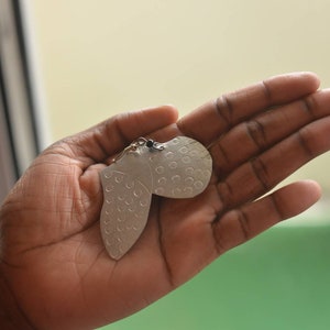 May include: A pair of silver earrings shaped like a heart with a textured surface. The earrings are held in a hand.
