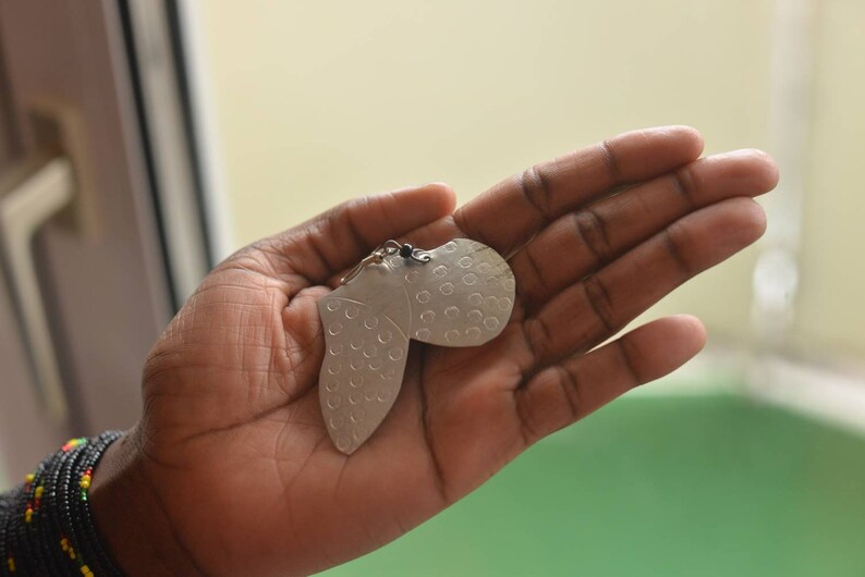 May include: A pair of silver earrings shaped like a heart with a textured surface. The earrings are held in a hand.