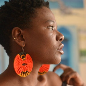 May include: A woman with short, dark hair wearing large, round earrings with a red, black, and white pattern. The earrings are made of fabric and have a floral design.