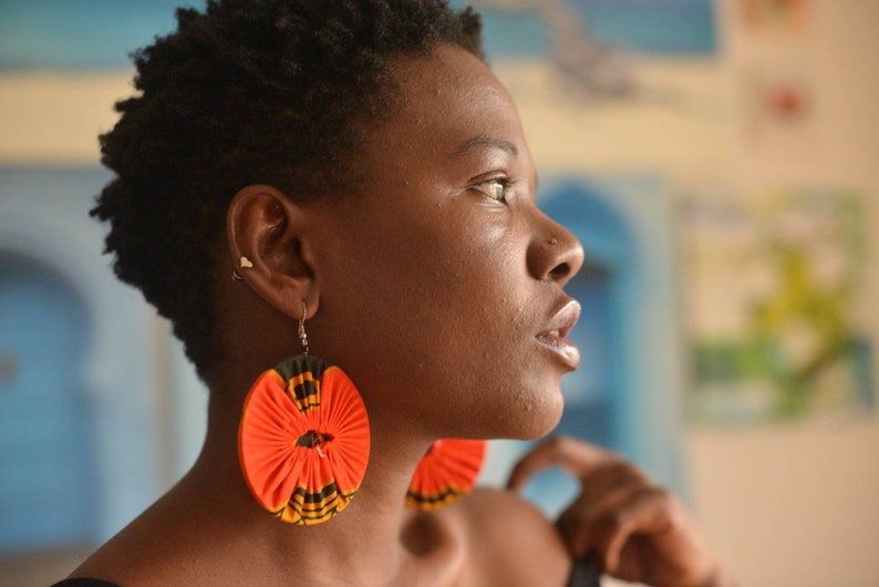 May include: A woman with short, dark hair wearing large, round earrings with a red, black, and white pattern. The earrings are made of fabric and have a floral design.