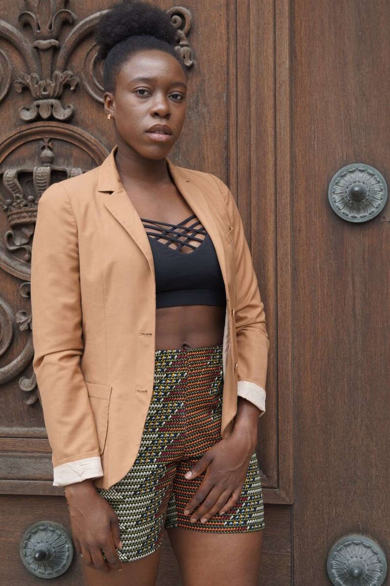 May include: A tan blazer with a black top and patterned shorts. The blazer has a classic design with a notched lapel and button closure. The shorts feature a colorful geometric pattern. The background is a wooden door.
