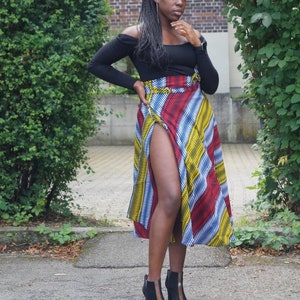 May include: A black off-the-shoulder top and a wrap skirt with red, yellow, blue, and white stripes. The outfit is completed with black ankle boots with a high heel. The skirt has a high slit. The outfit is worn outdoors.