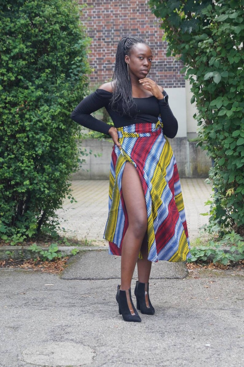 May include: A black off-the-shoulder top and a wrap skirt with red, yellow, blue, and white stripes. The outfit is completed with black ankle boots with a high heel. The skirt has a high slit. The outfit is worn outdoors.