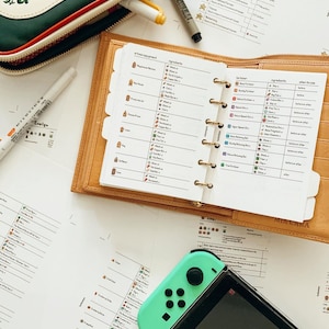 May include: A close-up of a brown leather planner with a white page open to a list of crafting recipes for the video game Animal Crossing: New Horizons. The planner includes a list of ingredients and when to use them for crafting various items. The planner is open to a page titled "Fertilizers" and lists the ingredients needed to make different types of fertilizer. The planner is lying on a white surface next to a Nintendo Switch console and a green controller.