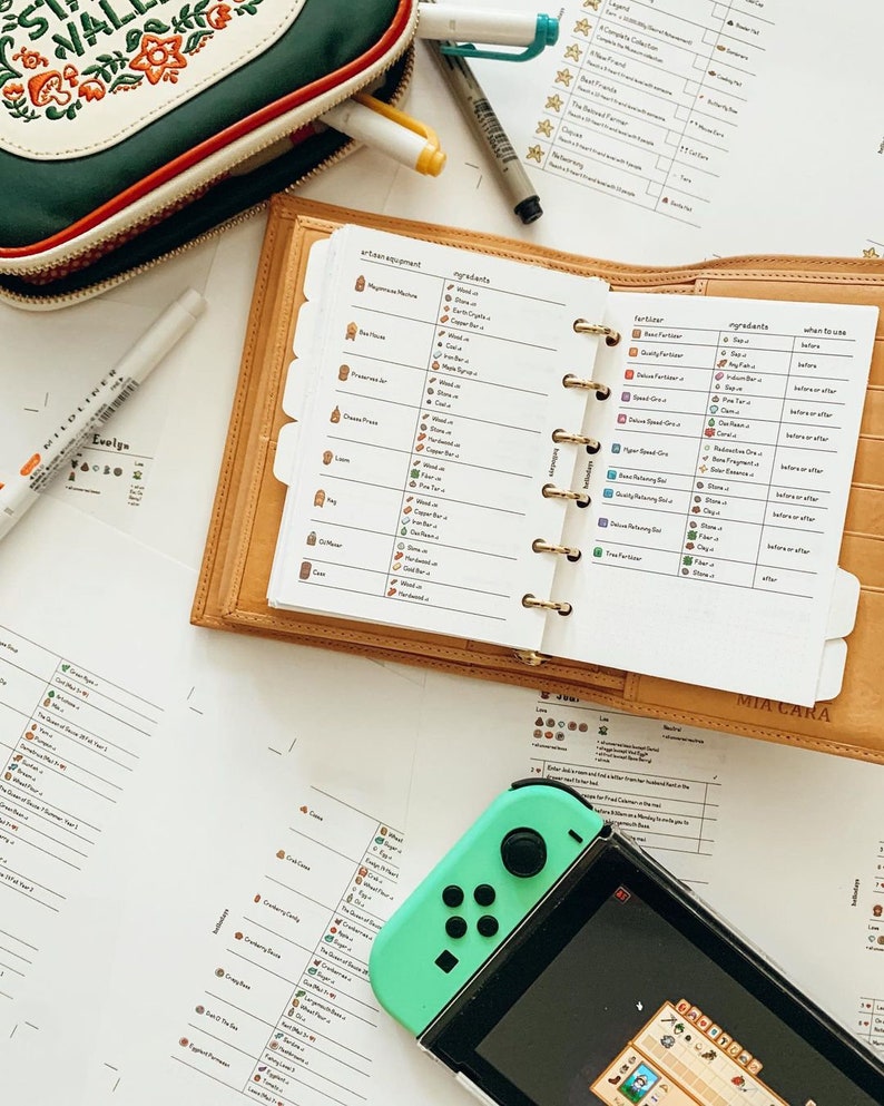 May include: A close-up of a brown leather planner with a white page open to a list of crafting recipes for the video game Animal Crossing: New Horizons. The planner includes a list of ingredients and when to use them for crafting various items. The planner is open to a page titled "Fertilizers" and lists the ingredients needed to make different types of fertilizer. The planner is lying on a white surface next to a Nintendo Switch console and a green controller.