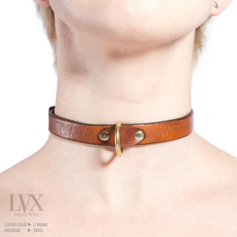 Pode incluir: Um colar de couro marrom com um anel de metal dourado.  LVX Supply & Co.  Cor do couro: Marrom claro.  Hardware: Lat&atilde;o.