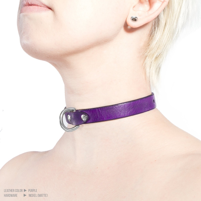 Pode incluir: Um colar de couro roxo com um anel de metal prateado. O colar &eacute; usado por uma pessoa com cabelo loiro.