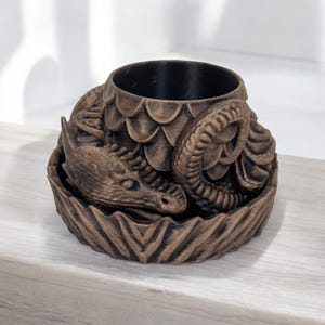 Pode incluir: Jardineira com tema de drag&atilde;o na cor bronze. A jardineira apresenta um design detalhado de drag&atilde;o, com a cabe&ccedil;a e o corpo do drag&atilde;o envolvendo o vaso cil&iacute;ndrico. A jardineira &eacute; feita de um material texturizado e tem um interior escuro.