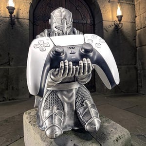 May include: A silver-coloured knight figurine kneeling and holding a white and black wireless video game controller. The knight wears detailed armour with chainmail textures and a helmet, presenting the controller forward with both hands.