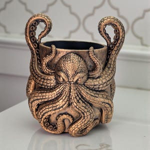 Puede incluir: Contenedor decorativo con temática de pulpo en color bronce. El contenedor presenta un diseño detallado de pulpo con tentáculos que forman asas. El interior es negro y el acabado tiene un brillo metálico. El contenedor probablemente esté hecho de resina o un material similar.