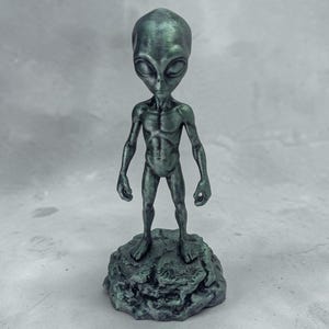 May include: A teal-colored alien figurine standing on a textured base. The alien has a large head, slender body, and large, oval eyes. The figurine is a decorative item, possibly for a desk or shelf.