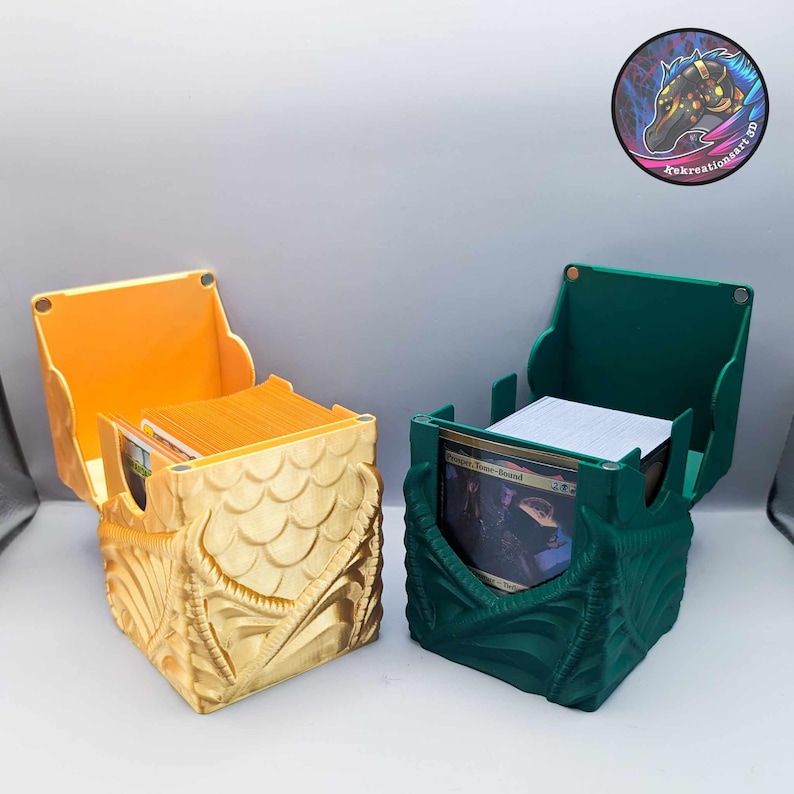 May include: Two 3D printed dragon-shaped card boxes, one gold and one green, with lids open and decks of cards inside. The boxes are designed with a dragon head and wings.