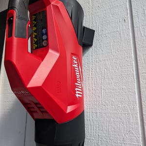May include: A red and black cordless leaf blower with the Milwaukee logo and FUEL text. The blower is mounted on a white wall.