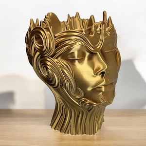 Pode incluir: Um vaso de planta de cor dourada, impresso em 3D, com a forma de uma cabeça humana com detalhes intrincados e uma expressão de olhos fechados.