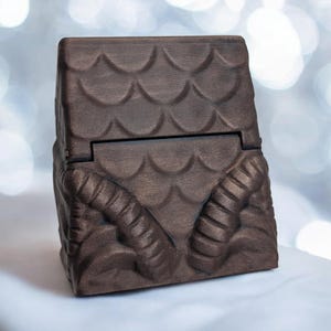 May include: A dark brown wooden box with a hinged lid. The box is decorated with a pattern of scales and two horns on the front.