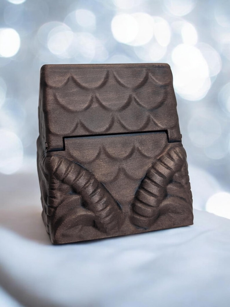 May include: A dark brown wooden box with a hinged lid. The box is decorated with a pattern of scales and two horns on the front.