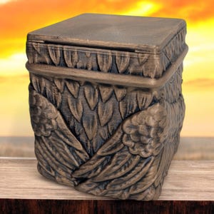 May include: A small, square, wooden box with a hinged lid. The box is decorated with a detailed carving of two angel wings. The box is painted in a bronze color.