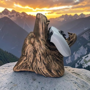 May include: A bronze-coloured wolf head sculpture with a textured fur finish, designed as a holder for a white video game controller. The wolf is depicted with its mouth open, providing a secure cradle for the controller to rest within its jaws.