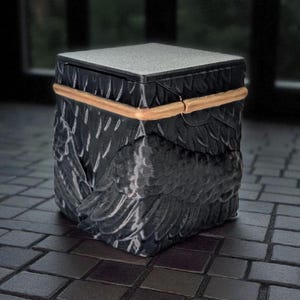 May include: A black, cube-shaped decorative box with a textured surface and a gold-colored band. The sides of the box are carved with a feather design, and the top is a light gray color. The box is sitting on a tiled surface.