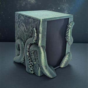 May include: A rectangular box with a metallic green finish featuring sculpted octopus tentacles wrapping around the sides. The surface has a textured, wood-like grain pattern, and the front panel is a smooth, dark, solid rectangular surface.