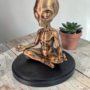 May include: A bronze-colored alien figure in a seated yoga pose, atop a black circular base. The alien has large oval eyes and is in a meditative position. A small potted succulent plant is in the background. The sculpture is on a wooden surface.