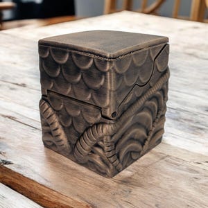 May include: A small, intricately carved wooden box with a bronze finish. The box has a hinged lid and a decorative design of scales and swirling patterns.