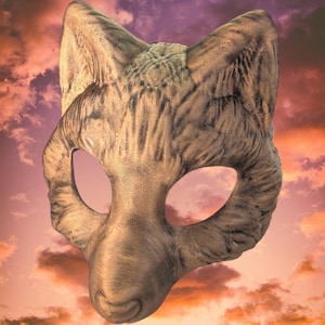 Op de afbeelding: Een gedetailleerd masker in de vorm van een wolvenkop, met een getextureerd oppervlak en licht- en donkerbruine markeringen. De oogopeningen zijn ovaal. De achtergrond toont een zonsondergang met oranje, roze en paarse kleuren.