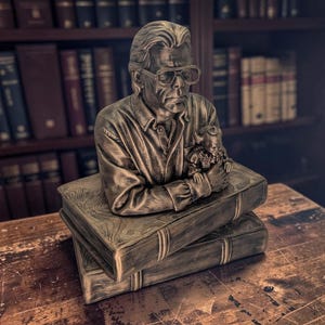 Peut inclure: Un buste couleur bronze représentant un homme à lunettes portant une chemise à col, tenant une petite figurine de clown. Le buste est posé sur deux livres rectangulaires empilés, dotés d'une finition texturée et vieillie.