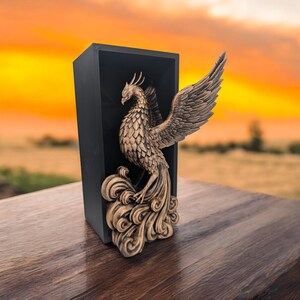 Könnte beinhalten: Eine goldfarbene Phönix-Figur mit ausgebreiteten Flügeln, die auf einer wellenförmigen Basis sitzt, wird in einer schwarzen Box präsentiert.
