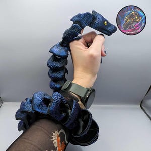 May include: A blue and black articulated dragon puppet with a segmented tail. The dragon has a textured, scaly body and a detailed head with yellow eyes.