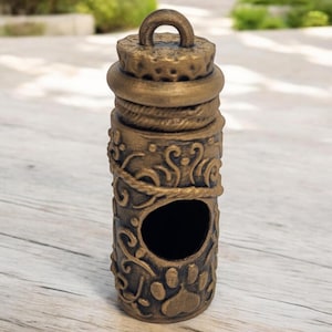 May include: A gold-colored, decorative, cylindrical pendant with a paw print design and a small opening. The pendant has a loop at the top for hanging.